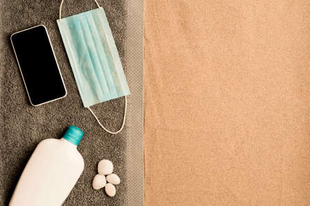 covid-19 prevention beach concept, cris towel on the beach sand, smartphone with black screen, blue virus protection mask, cream bottle and white stones. Sandy beach. Top view. space for textの写真素材