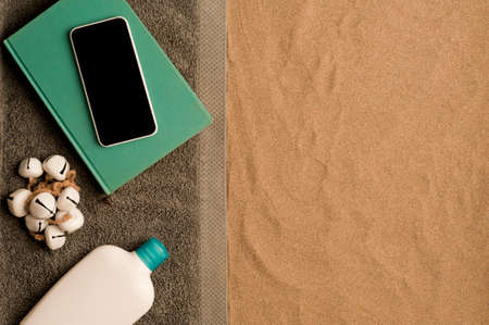 summer in the beach concept, cris towel on beach sand, on top of turquoise book with smartphone black screen, bottle of cream. beach sand .top view. space for textの写真素材