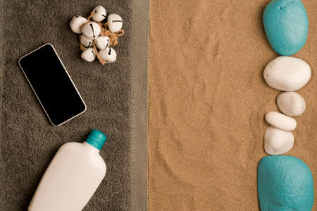 summer in the beach concept, cris towel on beach sand, black screen smartphone, bottle of cream and...