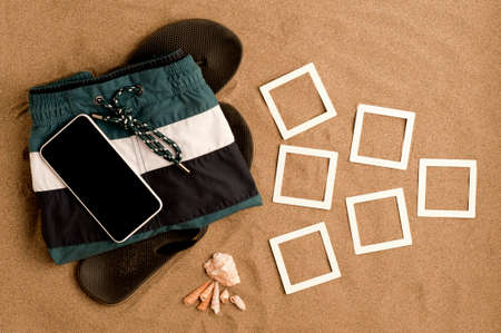 summer on the beach, black flip flops with trendy men's swimsuit and black screen smartphone, shells, frames mockup photo college on fine beach sand. Top view.の写真素材