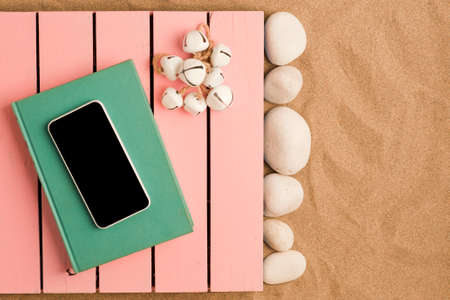 summer in the beach concept, pink wooden platform on beach sand, turquoise book with smartphone on...
