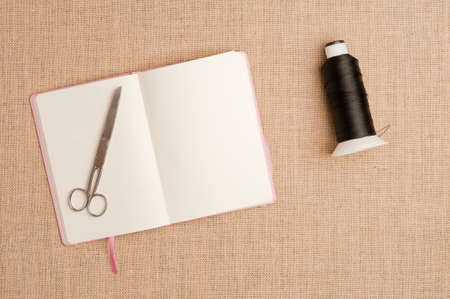 Texture or background of brown sackcloth and pink book with white sheets, scissors on top, sewing thread bobbin, empty space.の写真素材