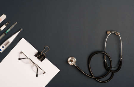Covid-19 Coronavirus concept. Stethoscope medicine accessories with notepad, eyeglasses and silver pen, thermometer, geringullia and coronavirus vaccine, black background with copy space. top viewの写真素材