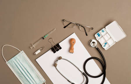 covid-19 concept (coronavirus) with medical instruments, protection material, (mask, stethoscope, vaccine syringe, pillbox with capsules, glasses and notepad), brown background, top viewの写真素材