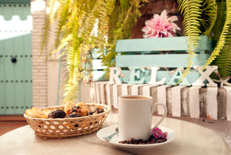 cup of coffee on marble table, (letters word relax), with pastries in wicker basket, and Mediterranean house background with plantsの写真素材