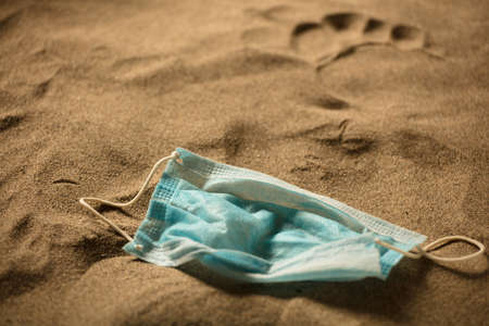 Waste during COVID-19. Discarded to single-use coronavirus face masks. Pollution of the environment, on the beach, man footprint in the sandの写真素材
