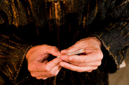 man hands with dark green djellaba, cigar in hand, prepares hashish cigar with white smoking paper,...