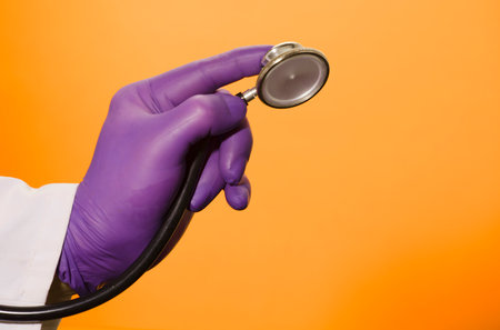 Stethoscope in doctor's hands with lilac surgical gloves, orange-yellow backgroundの写真素材