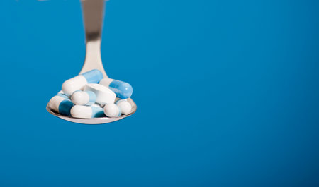 A group of pills on spoon with blue background. Blue and white pills in a spoon. Various pharmaceutical medicine pills, tablets and capsules in metal spoon. The pills are randomly distributedの写真素材