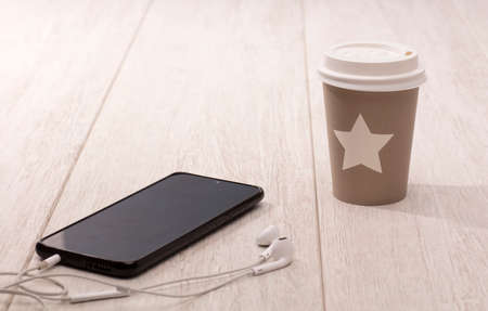 Disposable and recyclable tea cup with star, phone with headphones, on wooden table.