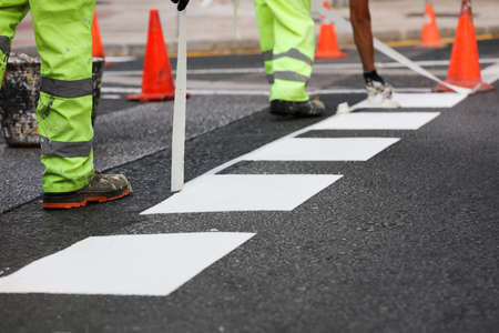 Painting the traffic line. Workers are painting white lines on the crosswalk. Road cones with orange and white stripes in the background, on asphalt during road construction worksの写真素材