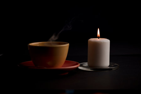 candle lit with flame, with cup of tea with smoke, black background. (focus on candle).の写真素材