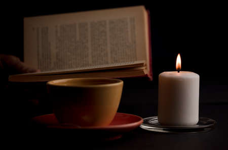 A candle lit with a flame, with a cup of tea with smoke, and a reading book in the background. (focus on candle).の写真素材