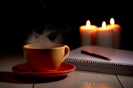 Cup of hot coffee with yellow smoke, notebook with pencil and burning candles. Desk table in power supply cutの写真素材