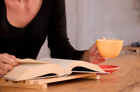 yellow cup with hot coffee, in woman's hands, with reading book at home. Focus on the cup.の写真素材