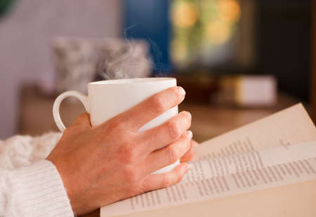 white cup with hot drink and smoke, hands warming up from the heat, at home. Focus on the cupの写真素材