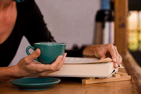 green cup with hot coffee, in woman's hands, with reading book at home. Focus on the cupの写真素材