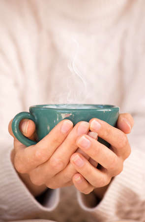 Close up of female hands with a cup of drinks. Beautiful girl in white sweater holding a green cup of tea or coffee in the morning sunlight. Focus on the cup.の写真素材