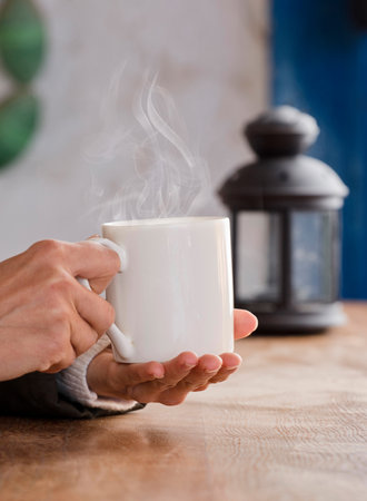 white cup with hot drink and smoke, hands warming up from the heat, at home. Focus on the cup.の写真素材