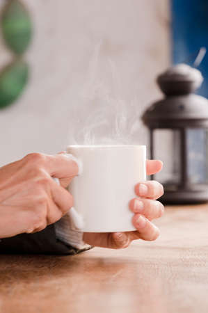 white cup with hot drink and smoke, hands warming up from the heat, at home. Focus on the cup.の写真素材