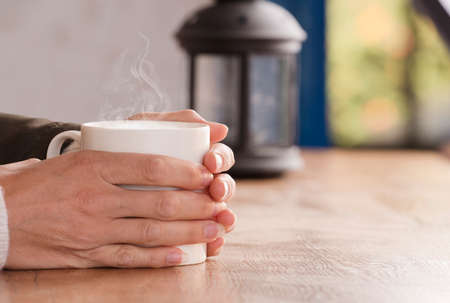 white cup with hot drink and smoke, hands warming up from the heat, at home. Focus on the cup.の写真素材