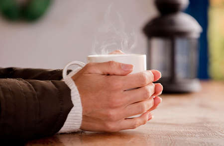 white cup with hot drink and smoke, hands warming up from the heat, at home. Focus on the cup.の写真素材