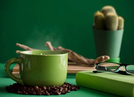 cup of hot green coffee with steam, old wooden trunk, cactus, book and reading glasses, space for text, dark green background.の写真素材