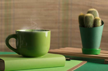 cup of tea or coffee, green color, hot with steam, book and cactus, with table and light brown background.の写真素材