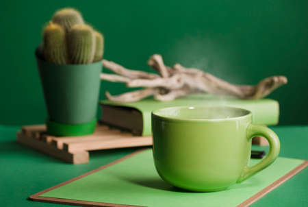 cup of hot green coffee with steam, old wooden trunk, cactus, book and reading glasses, space for text, dark green background.の写真素材