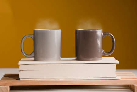 Two brown and gray cups of coffee or tea with hot steam on the table, with book and brown background.の写真素材