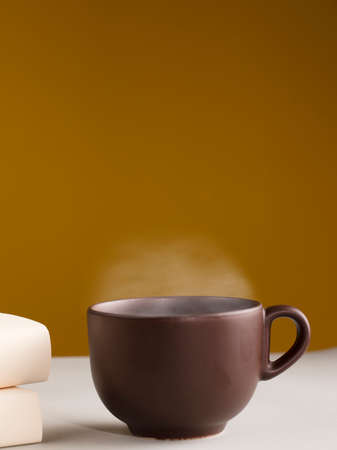 Brown cup of coffee or tea with hot steam, on the table, with books, glasses and brown background.の写真素材