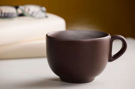 Brown cup of coffee or tea with hot steam, on the table, with books, glasses and brown background.の写真素材