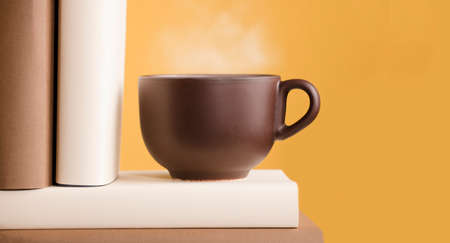 Brown cup of coffee or tea with hot steam, on the table, with booksの写真素材
