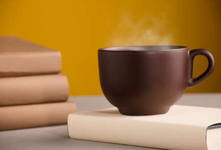 Brown cup of coffee or tea with hot steam, on the table, with booksの写真素材