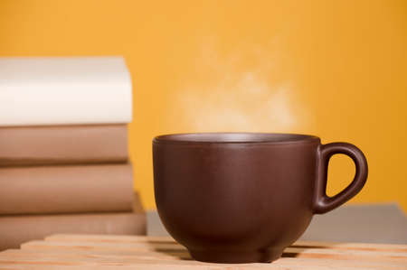 Brown cup of coffee or tea with hot steam, on the table, with booksの写真素材