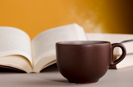 desk with cup of coffee or tea with hot steam, open books, orange yellow backgroundの写真素材