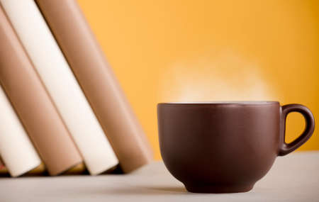 Brown cup of coffee or tea with hot steam, on the table, with booksの写真素材