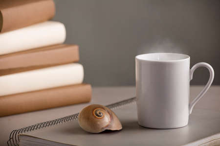 white cup of coffee with hot steam, on the desk with nautilus shell and books, gray backgroundの写真素材