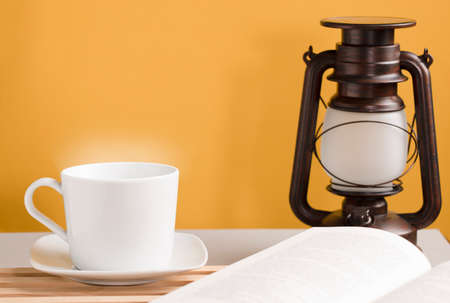 white cup of coffee with hot steam, on the desk with open book and sunlight lamp, orange-yellow backgroundの写真素材