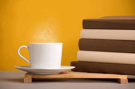 white cup of coffee with hot steam, on the desk with brown books, yellowish orange backgroundの写真素材