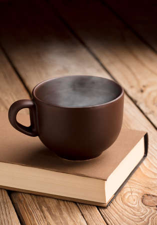 Brown cup of coffee with smoke on reading book, rustic wooden table, at sunrise.の写真素材