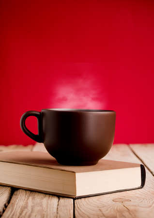 Brown coffee cup with smoke on reading book, rustic wooden table, red background.の写真素材