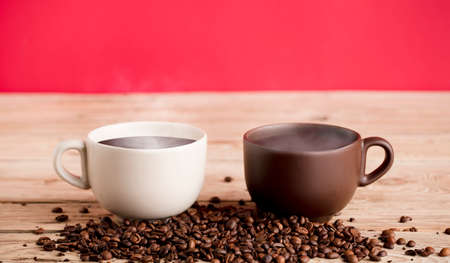 Two brown and off white coffee cups with smoke and beans on rustic wooden table, red background.の写真素材