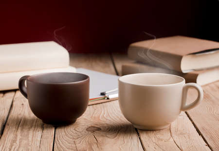 brown cup and white cup with chocolate, tea or coffee, hot steam, books on rustic wooden table with red backgroundの写真素材
