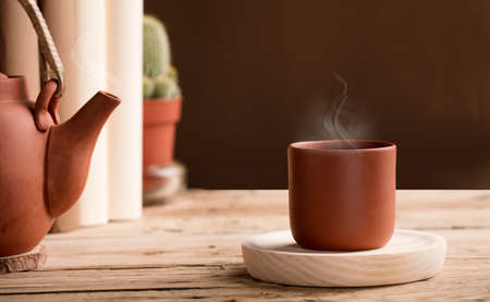 Cup of tea or coffee with hot steam from brown clay, on wooden table with partial view of teapot or coffee pot. books, cactus.の写真素材
