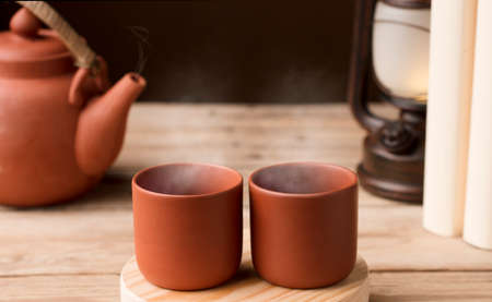 Two cups with hot steamed brown clay tea or coffee, on a wooden table with partial view of the lamp and the kettle or coffee pot. brown background. Space for textの写真素材