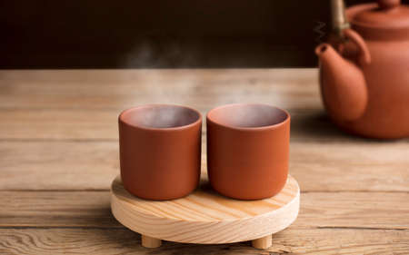 Two cups with hot steamed brown clay tea or coffee, on a wooden table with partial view of the kettle or coffee pot. brown background. Space for text.の写真素材