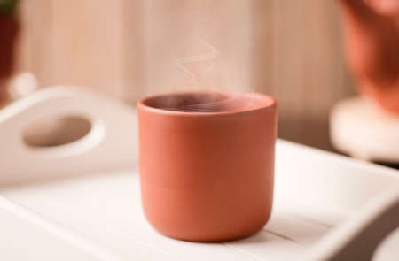 Earthenware mug with hot steam, coffee or tea on a white wooden tray, with partial view of the kettle, brown wooden background.の写真素材