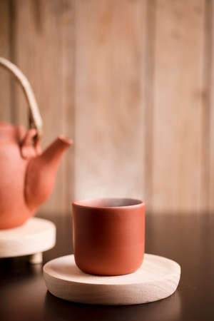 Earthenware mug with hot steam, coffee or tea on wooden table, with partial view of kettle, brown wooden background.の写真素材
