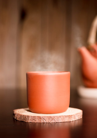 Earthenware mug with hot steam, coffee or tea on wooden table, with partial view of kettle, brown wooden background.の写真素材